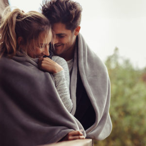 Romantic couple on a winter holiday. Man and woman standing together in a hotel room balcony wrapped in blanket. Couple embracing and smiling.